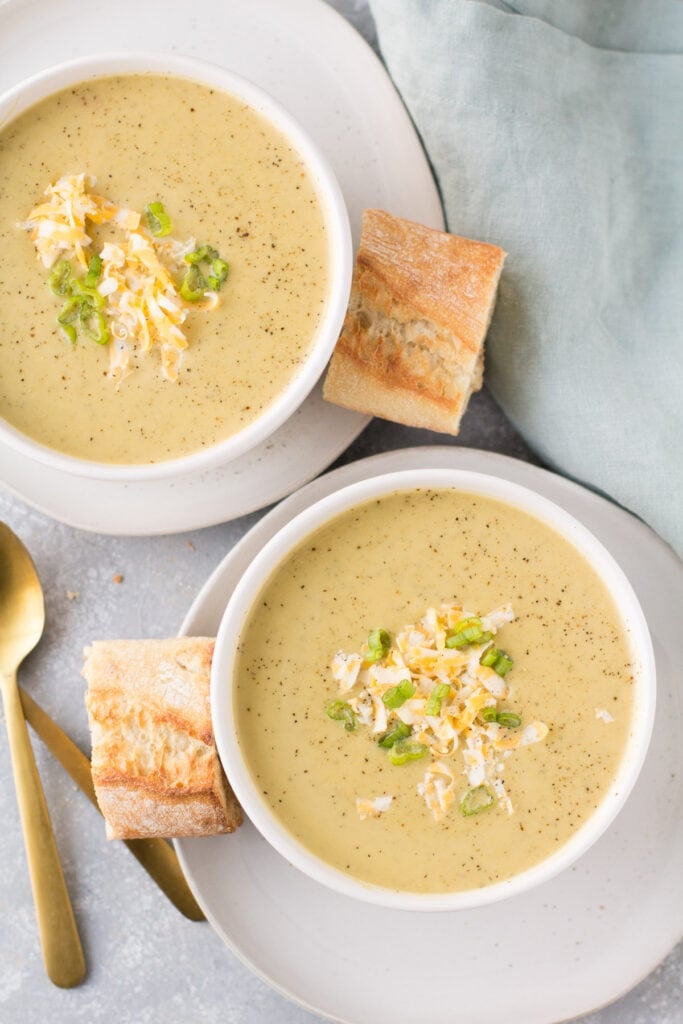 An overhead shot of 2 bowls of healthy broccoli soup with bread and blue linen off to the side