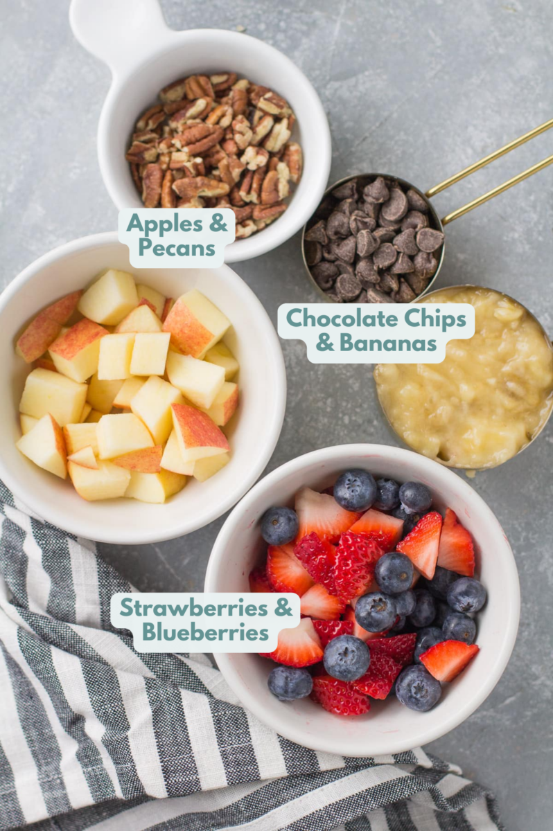 Toppings for baked steel cut oatmeal in small white bowls and measuring cups: apple, chopped pecans, chocolate, mashed banana, fresh berries (fresh blueberries and strawberries)