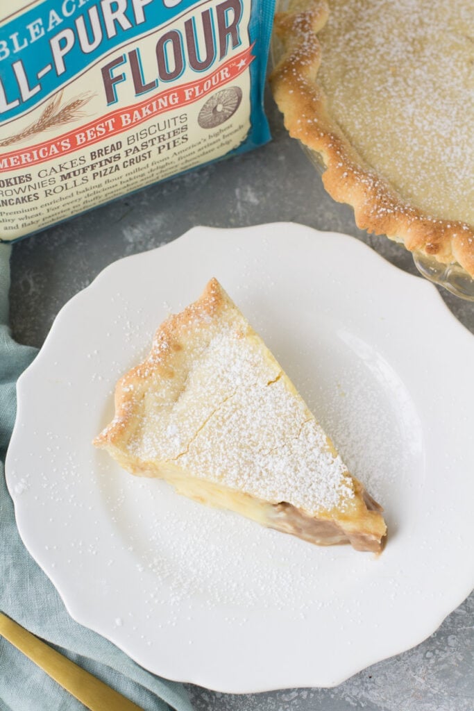 A slice of Italian Cream Pie ona white plate with a pie off to the side and a bag of flour off to the other side