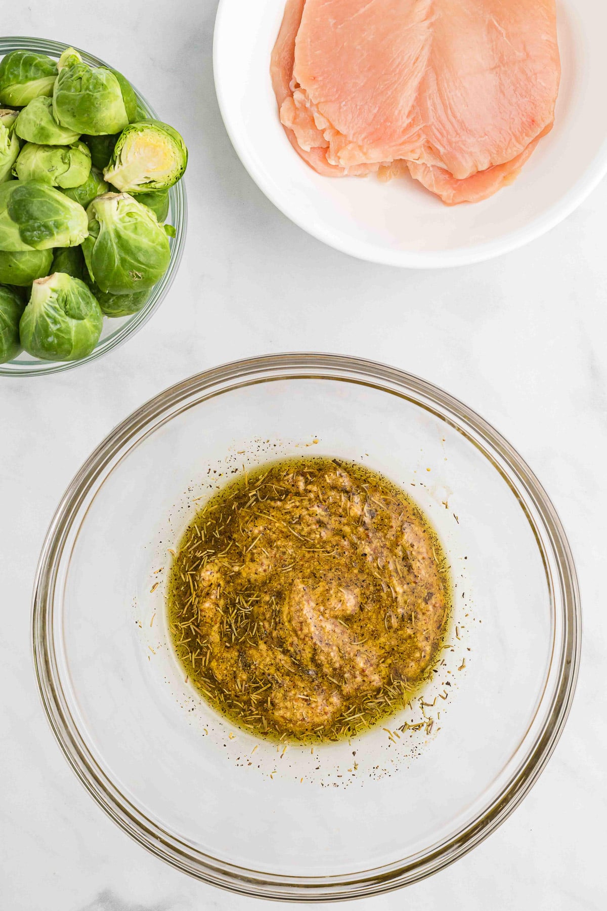 Maple mustard chicken marinade in a clear large bowl with chicken and brussels sprouts on the side