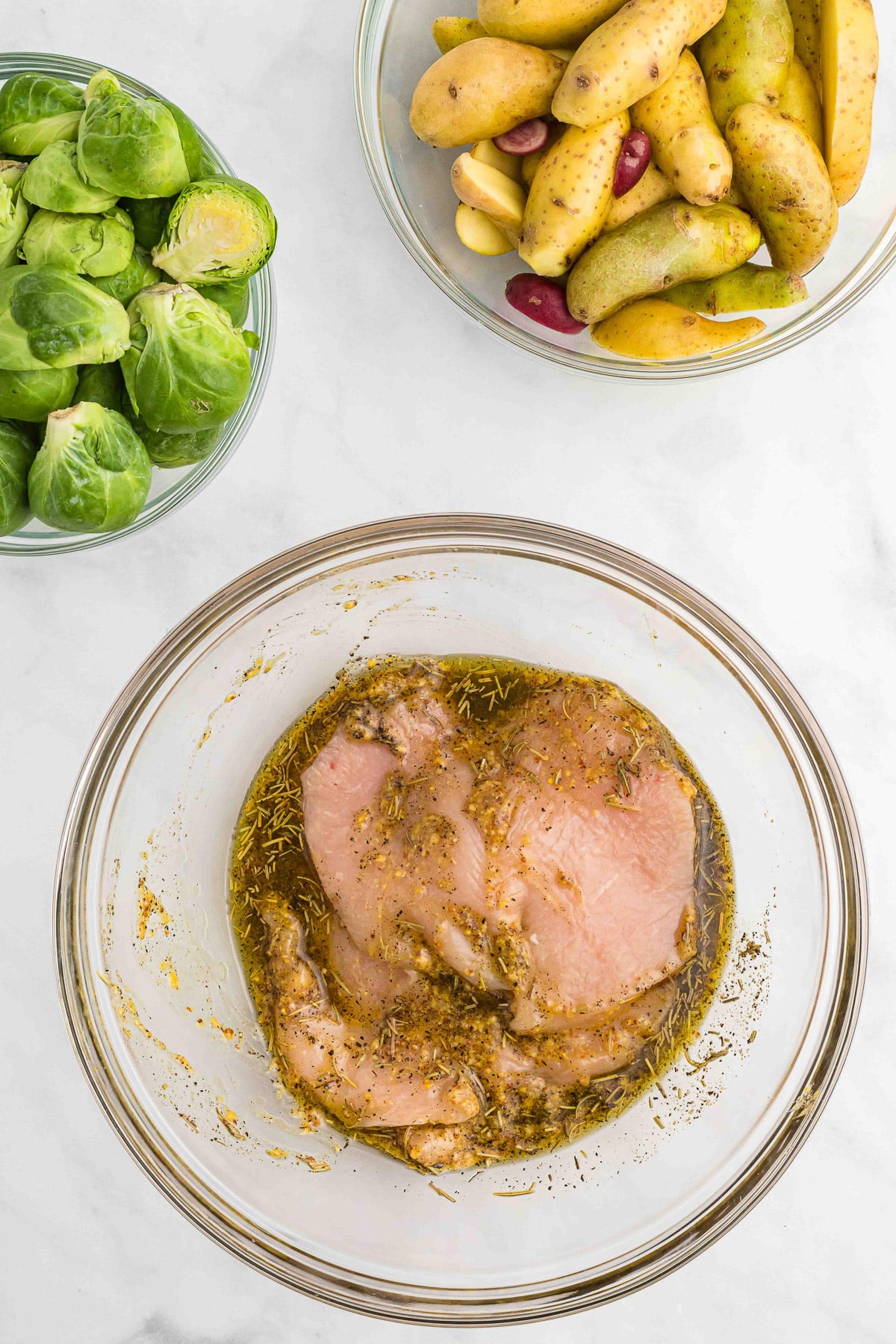 chicken breasts marinating in maple mustard chicken pieces marinade in a clear large bowl with brussels sprouts and potatoes in a small bowl on the side