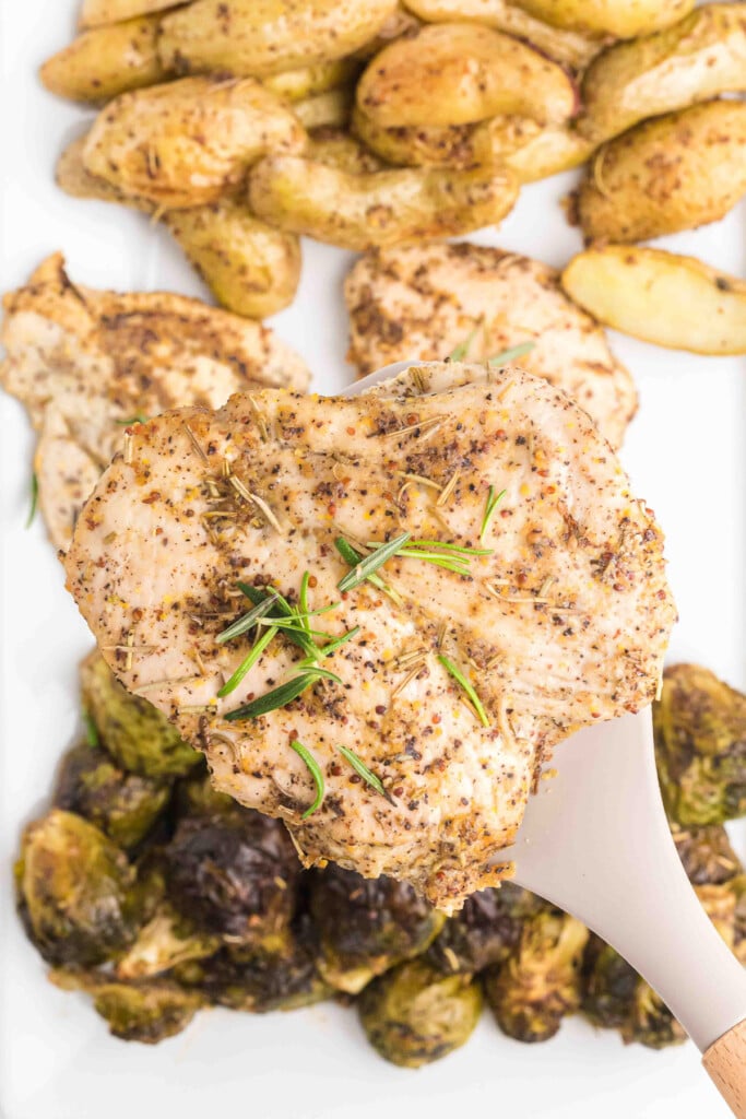 a maple mustard chicken breast being lifted off a platter of chicken, brussels sprouts, and potatoes 