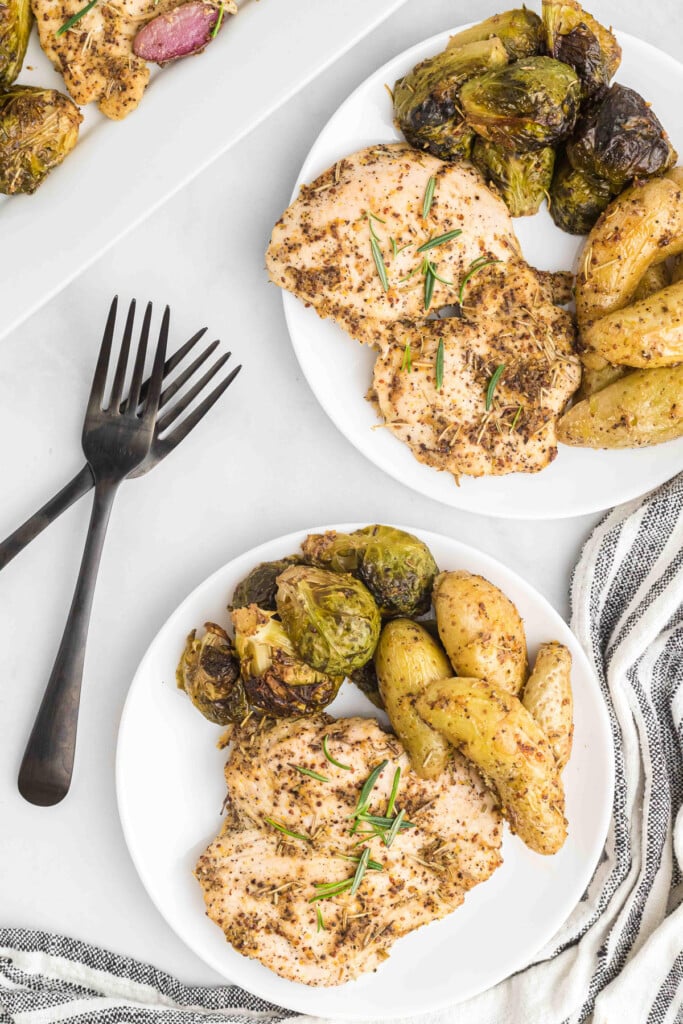 two white plates of maple mustard chicken breast, brussels sprouts and potatoes with forks on the side on a white background