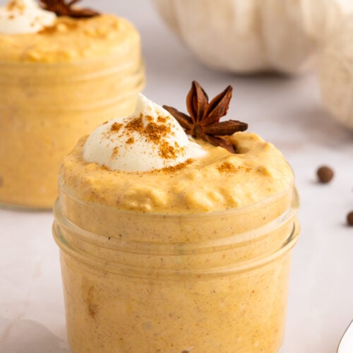 Easy Pumpkin Overnight Oats Recipe in two small clear mason jars topped with cream, pumpkin spice, and a star anise.