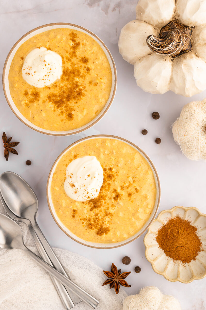 An overhead shot of two bowls of pumpkin overnight oats topped with a dollop of whipped cream and pumpkin pie spice and spices on the side.