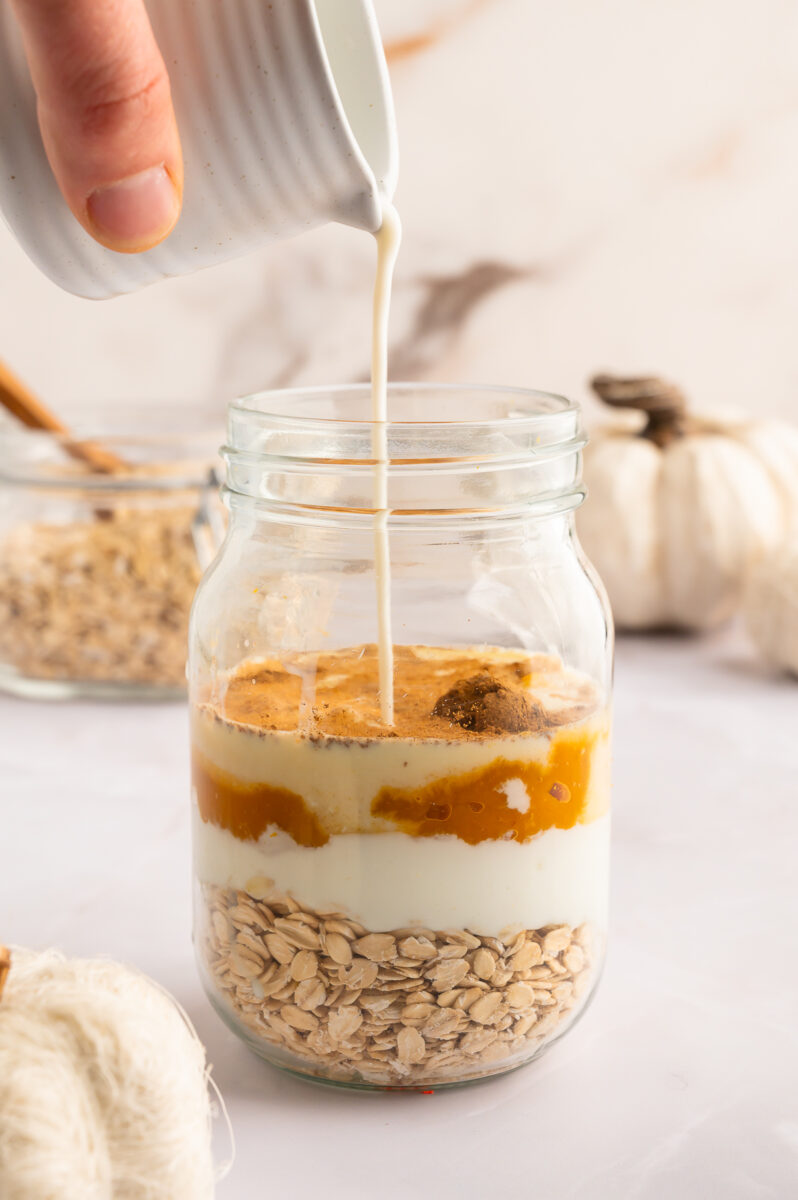 Milk being poured into a glass mason jar with ingredients for pumpkin overnight oats layered in the jar.