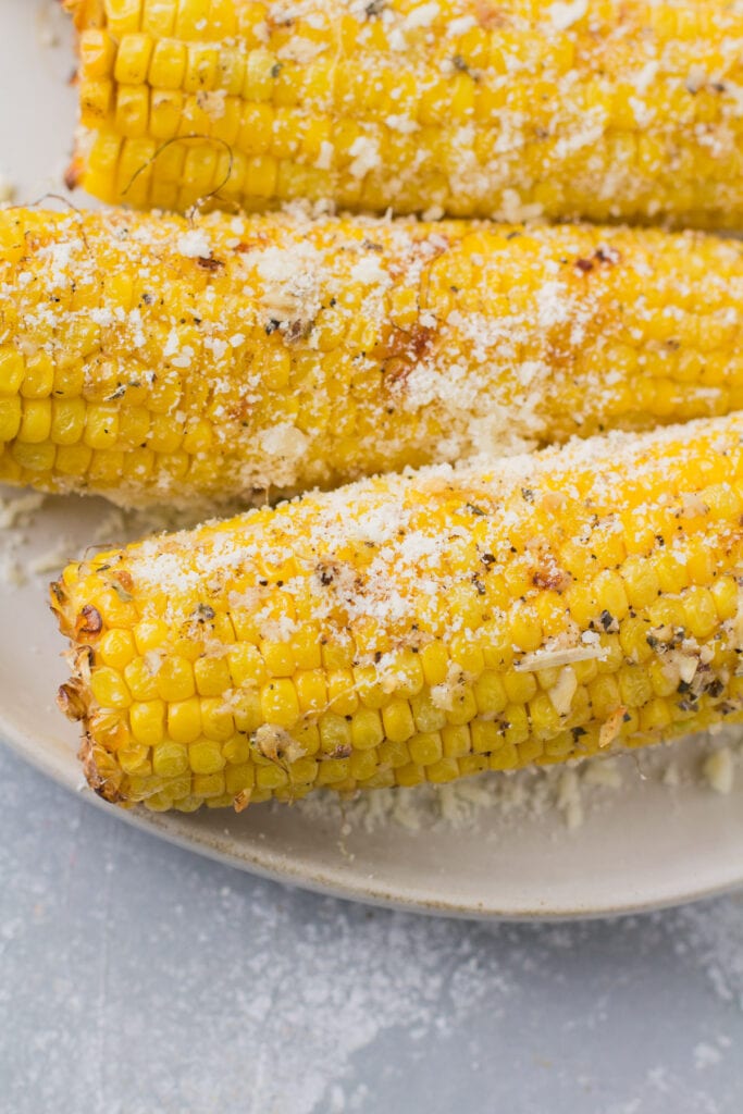 A close up of garlic parmesan grilled corn on a white plate on a grey background