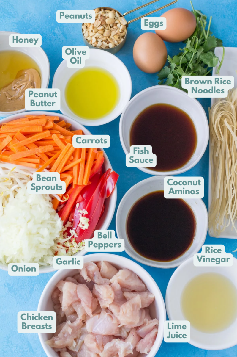 Ingredients for Healthy Pad Thai in small white bowls on a bright blue background - veggies, honey, oil, fish sauce, coconut aminos, rice wine vinegars, peanuts, noodles and chicken.