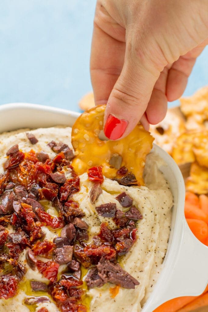 A hand with a pretzel chip scooping white bean hummus out of a dish