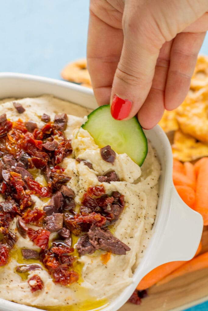 A hand dipping a slice of cucumber into vegan white bean hummus