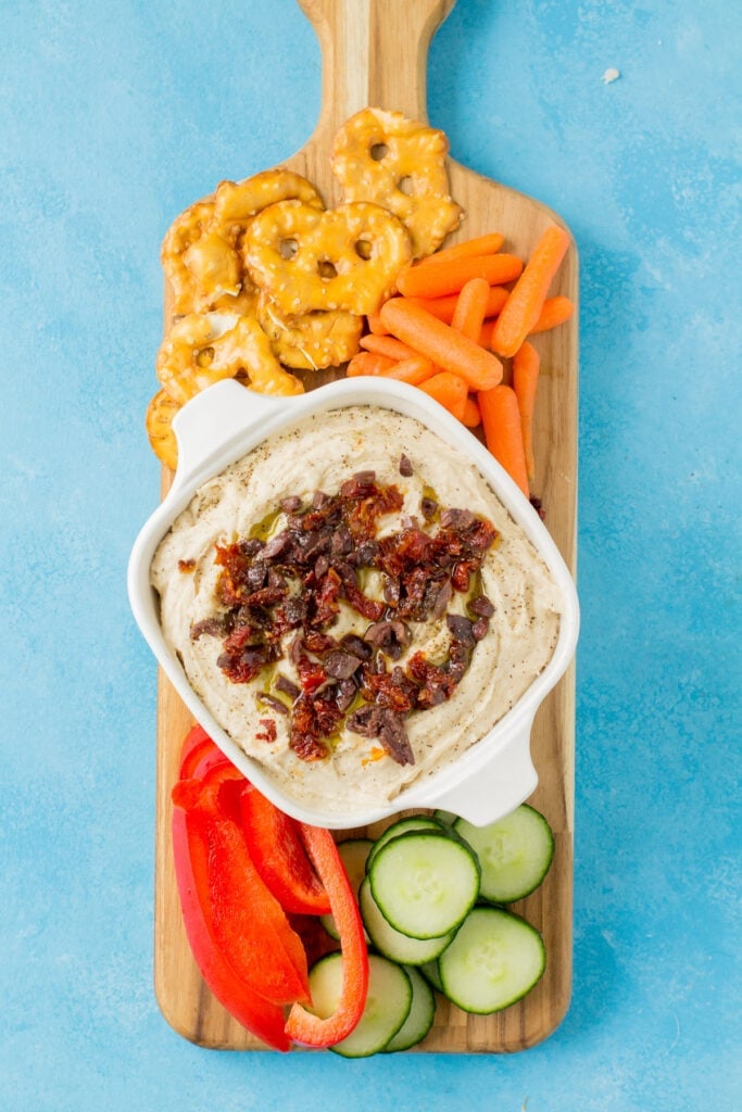 White bean hummus on a wooden cutting board with peppers, carrots and cucumbers off to the side on a bright blue background