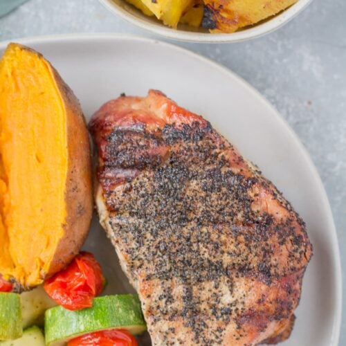 A close up of a grilled pork chop on a white plate with veggies, a potato and pineapple