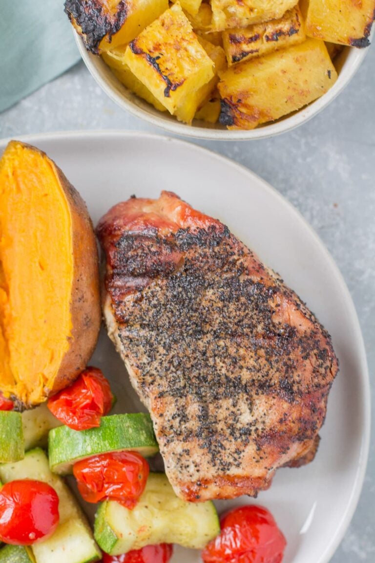 A close up of a grilled pork chop on a white plate with veggies, a potato and pineapple