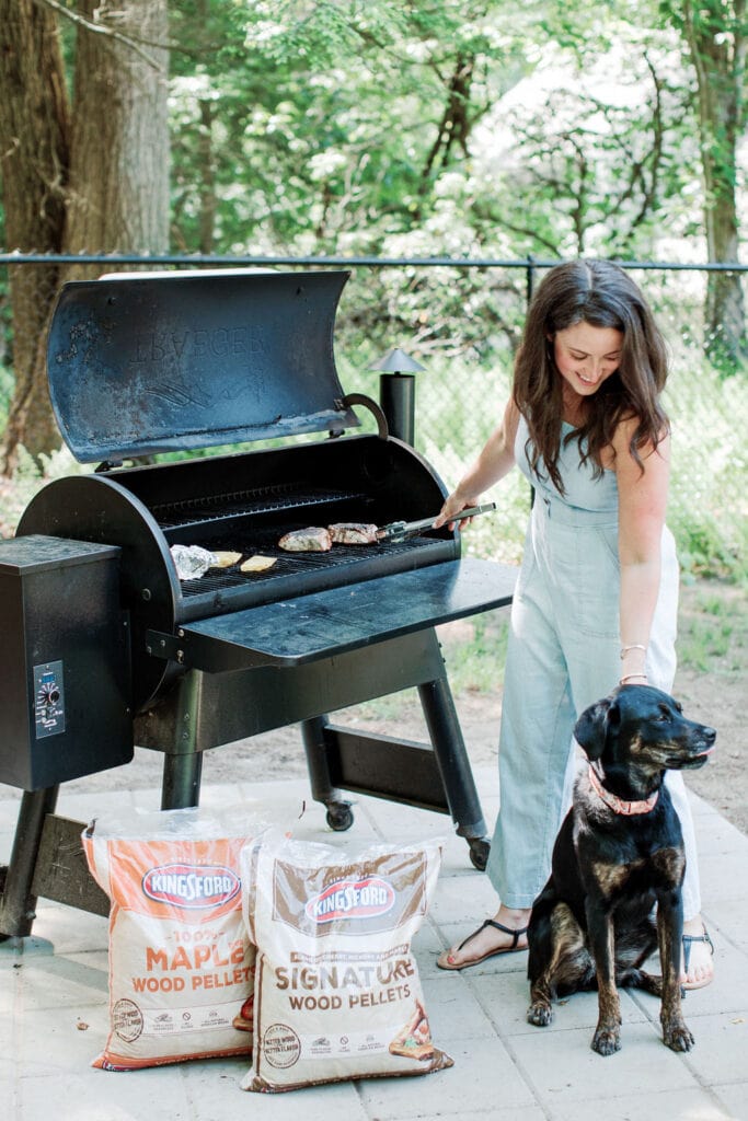 A girl standing at a grill with kingsford pellets off to the side.