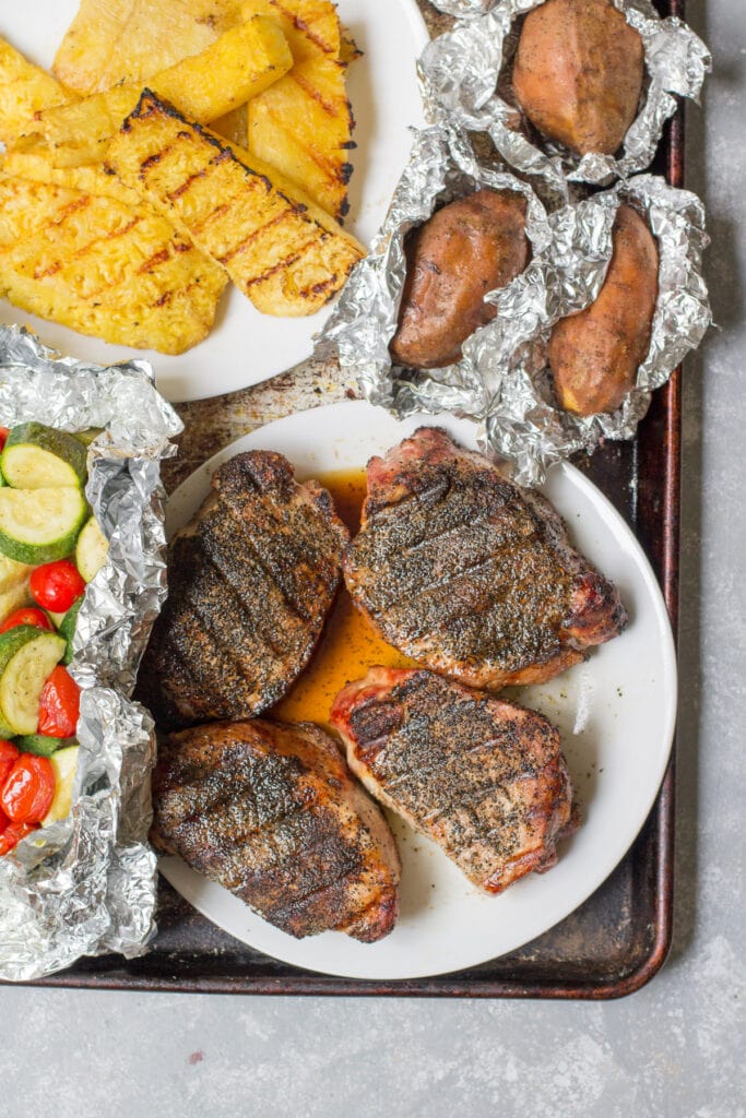 An overhead shot of a grilled dinner on a baking sheet - pork chops, veggies, potatoes and pineapple