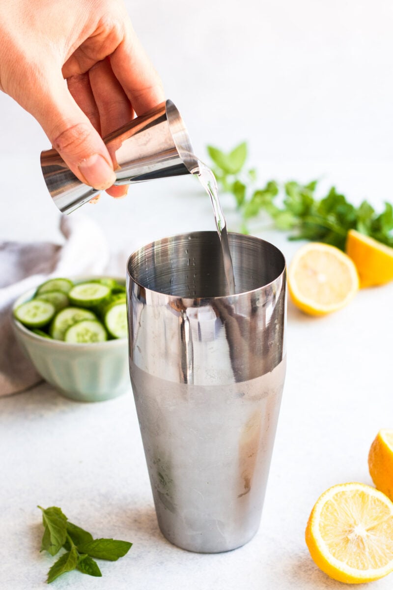 A hand pouring vodka into a cocktail shaker with lemons and cucumbers in the background.
