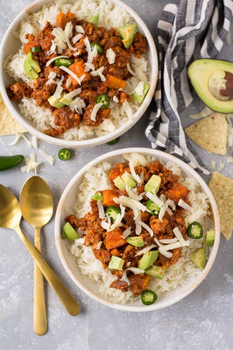 Ground Beef and Sweet Potato Chili in small white bowls with jalapenos, gold spoons, avocados and chips off to the sides