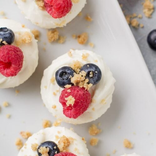 An overhead shot of mini healthy cheesecakes on a white square plate with berries and crumbs off to the side