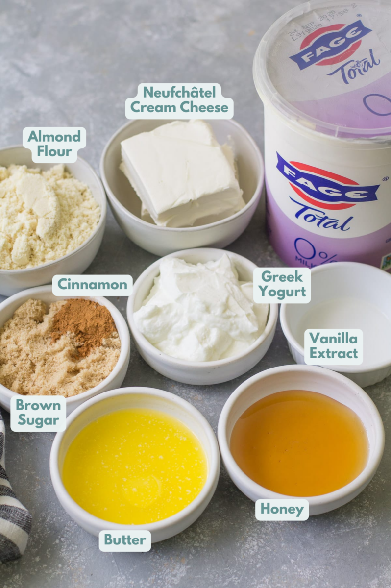 Ingredients for healthy cheesecakes in small white bowls: almond flour, cream cheese, yogurt, vanilla extract, honey, butter, brown sugar and cinnamon