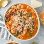A close up of a bowl of easy minestrone soup with cheese, spoons and dried herbs off to the side.
