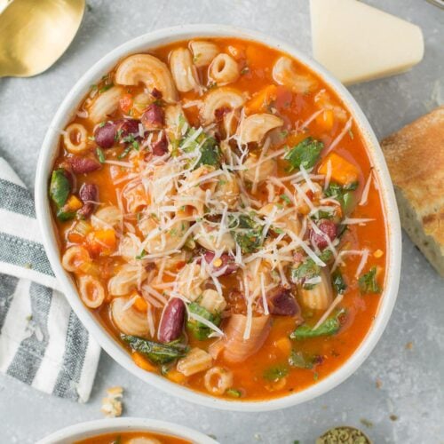 A close up of a bowl of easy minestrone soup with cheese, spoons and dried herbs off to the side.