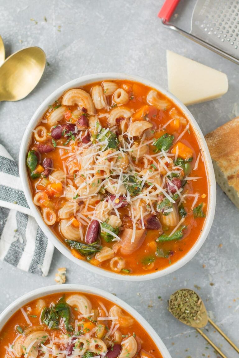 A close up of a bowl of easy minestrone soup with cheese, spoons and dried herbs off to the side.