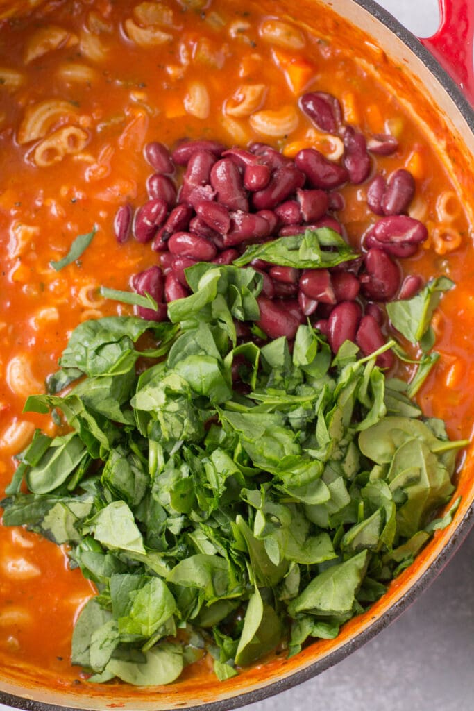 a close up of minestrone soup with spinach and beans being added to it.