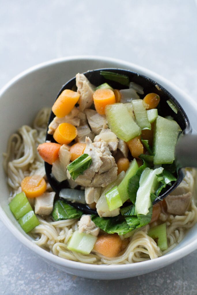 a ladle adding Asian Chicken Soup to a bowl of cooked ramen noodles.