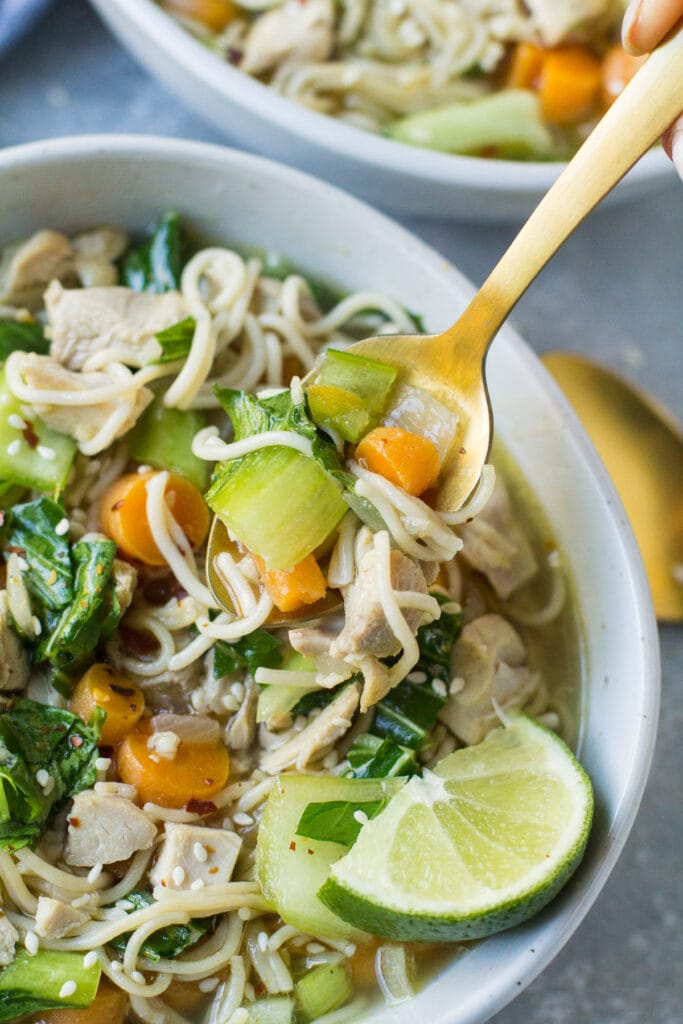 A spoon taking a scoop out of a bowl of loaded Asian Chicken Soup - veggies, noodles and a lime wedge fill the bowl