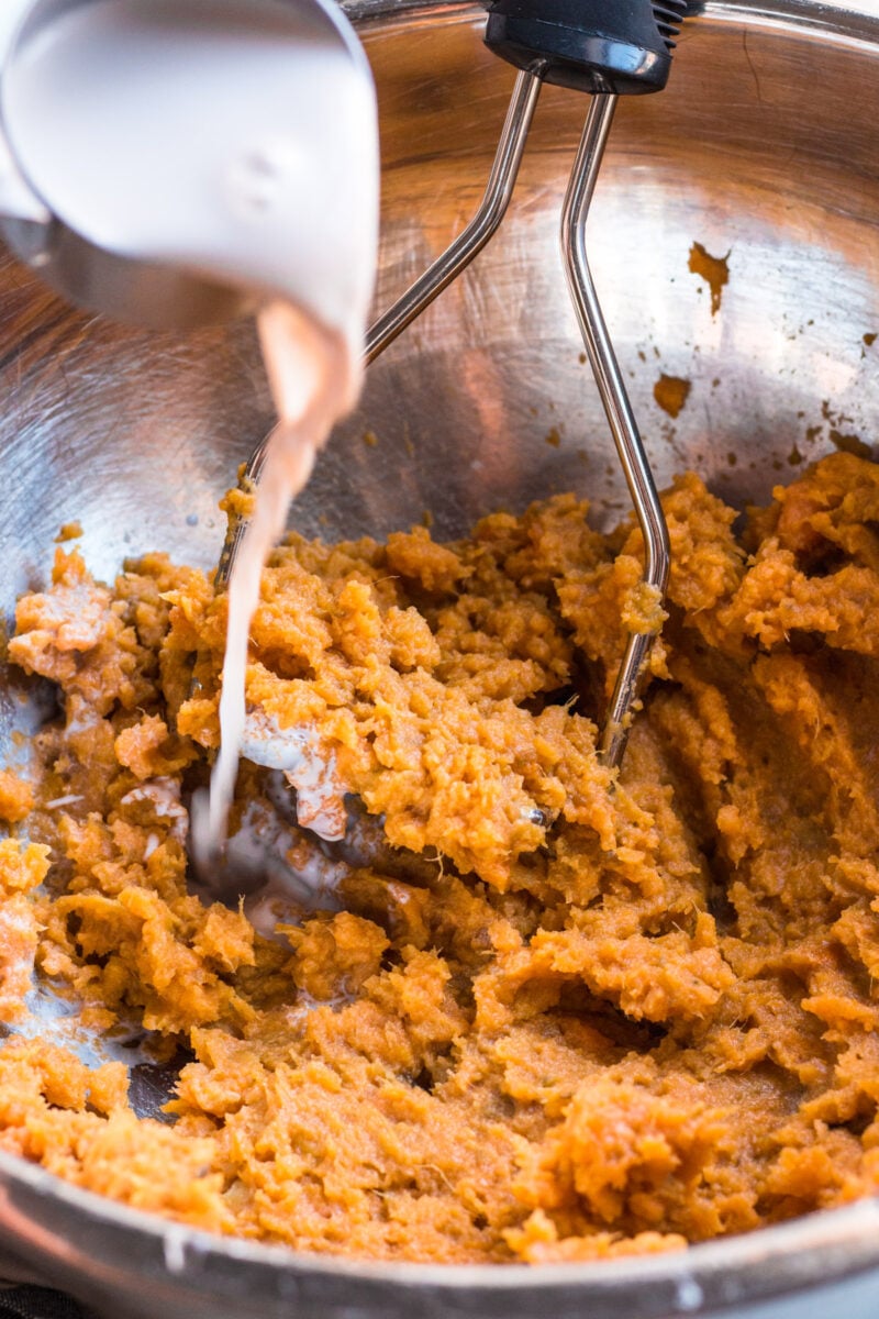 Mashed sweet potatoes in a bowl with a masher in it with milk being poured in. 