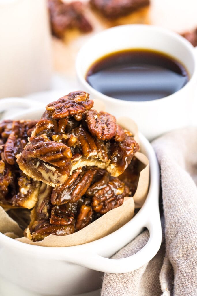A close up of gluten free pecan pie bars in a small white bowl with a cup of coffee in the background.