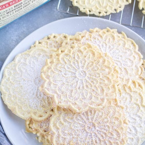 Pizzelle Cookies on a white plate with a cooling rack and back of flour off to the side.
