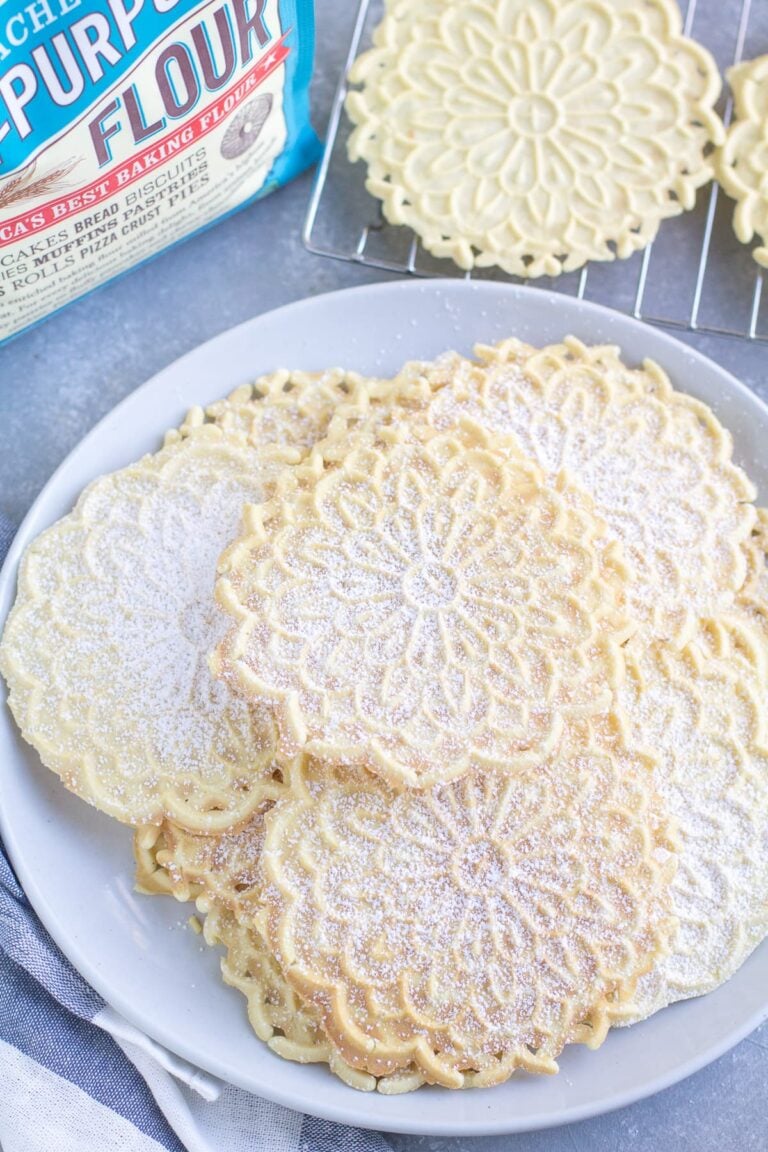 Pizzelle Cookies on a white plate with a cooling rack and back of flour off to the side.