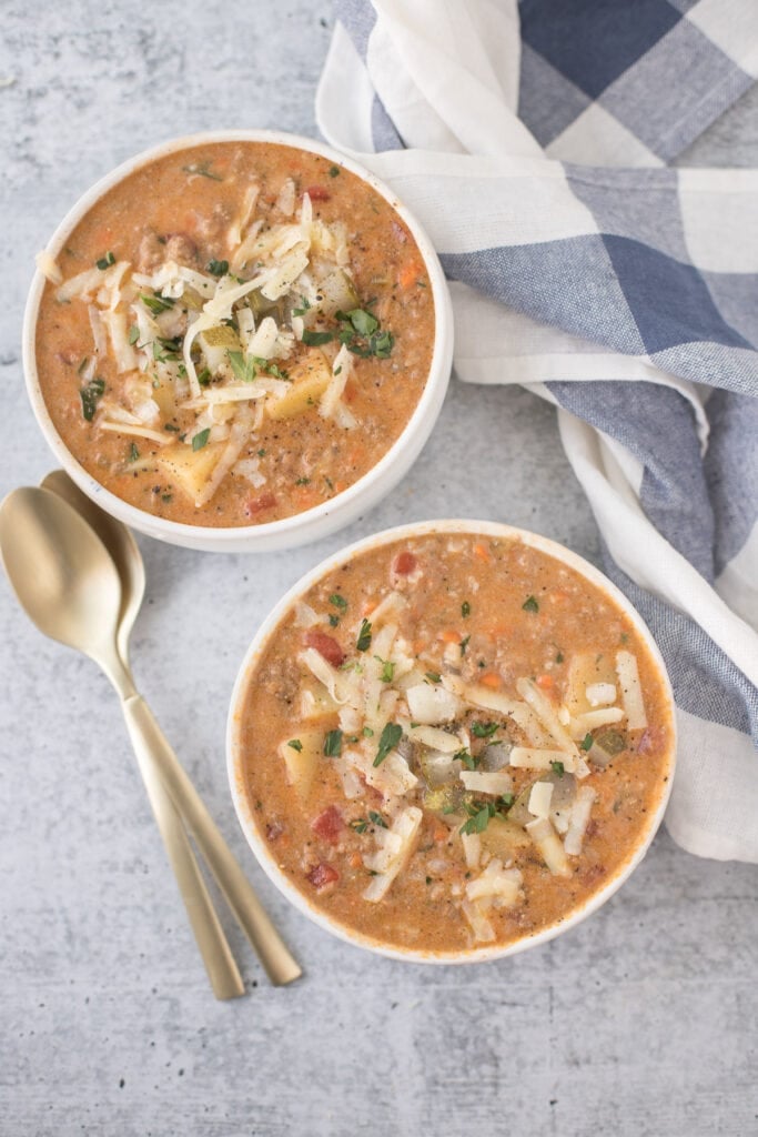 Two bowls of crockpot cheeseburger soup on a grey background with a blue checked linen off to the side