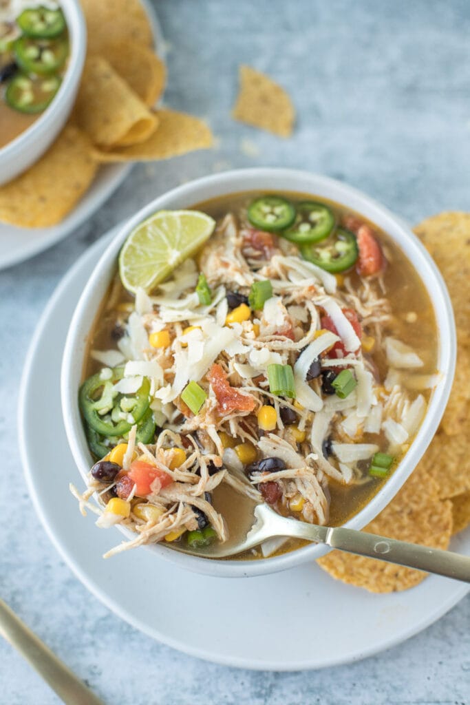 Chicken tortilla soup in a white bowl with a spoon dipped in and with a side of chips on a grey background