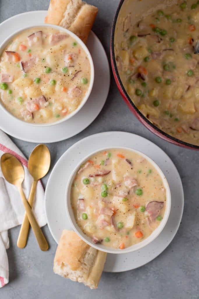 Ham and potato soup in white bowls with bread on the side on a grey background with a pot of ham and potato soup in the corner and gold spoons