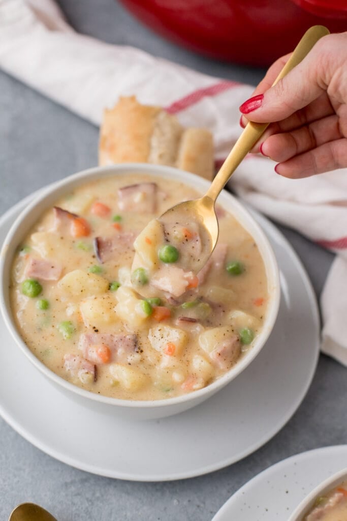 A white bowl of ham and potato soup with a gold spoon taking a spoonful out with bread on the side and a white and red towel and red pot in the background