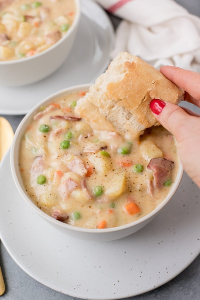 A white bowl filled with ham and potato soup with a piece of bread being dipped into it on a grey background with another bowl of soup on the side