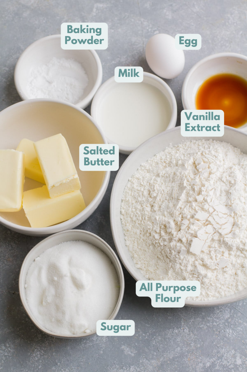 Cut out sugar cookie ingredients - butter, flour, sugar, milk, vanilla, an egg and baking powder in small white bowls