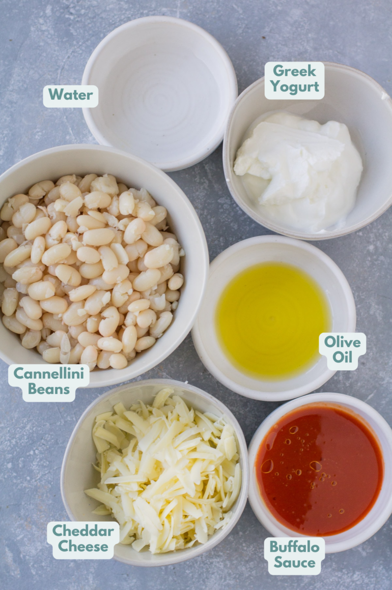 Ingredients for Vegetarian Buffalo Dip in small white bowls on a grey background: water, nonfat greek yogurt, extra virgin olive oil, buffalo sauce, cheddar cheese, cooked cannellini beans