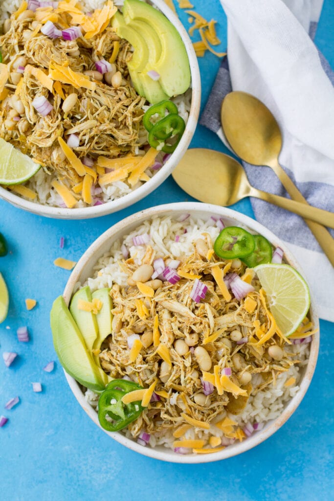 Top view of two bowls of healthy white chicken chili in white bowls with two gold spoons on the side and a white and blue striped dish towel on the side over a bright blue background
