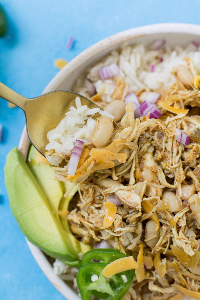 A top view close up of healthy chicken chili in a white bowl with a gold spoon taking a bite out on a bright blue background