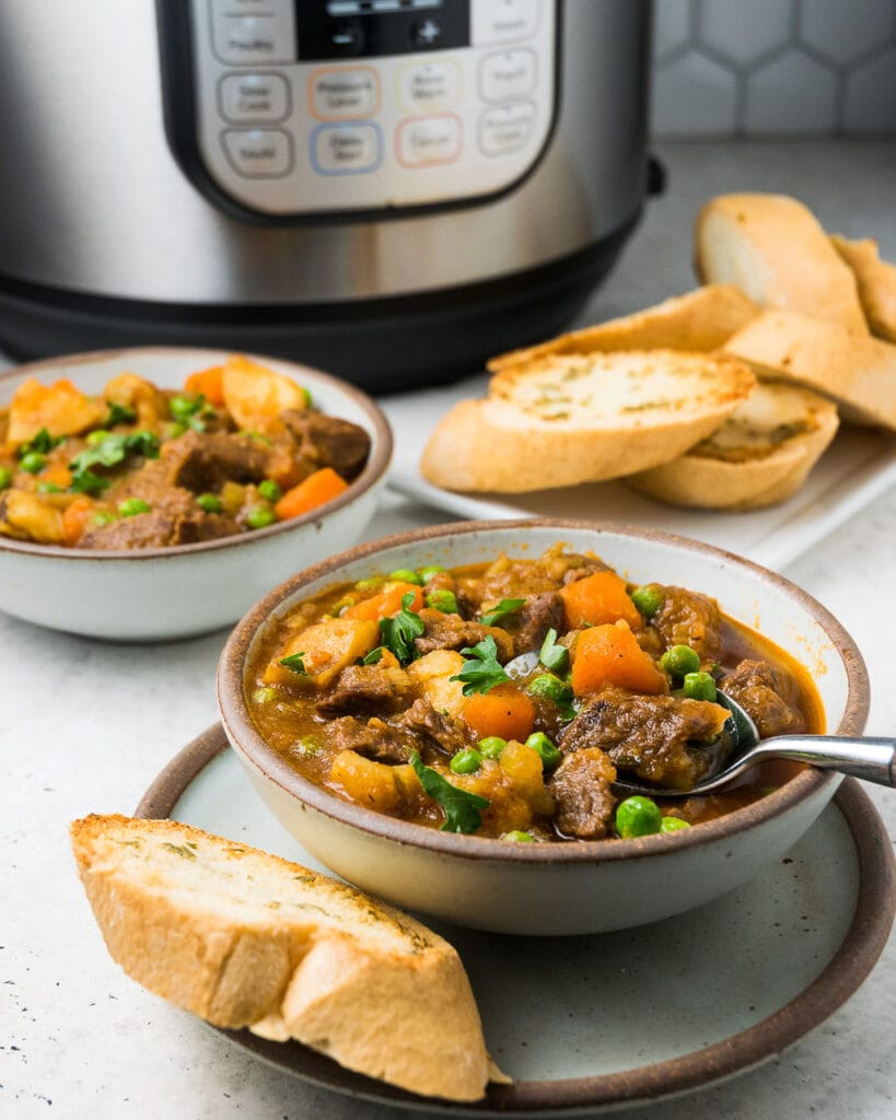 Two white bowls with beef stew with bread on the side on a white background with an instant pot in the back