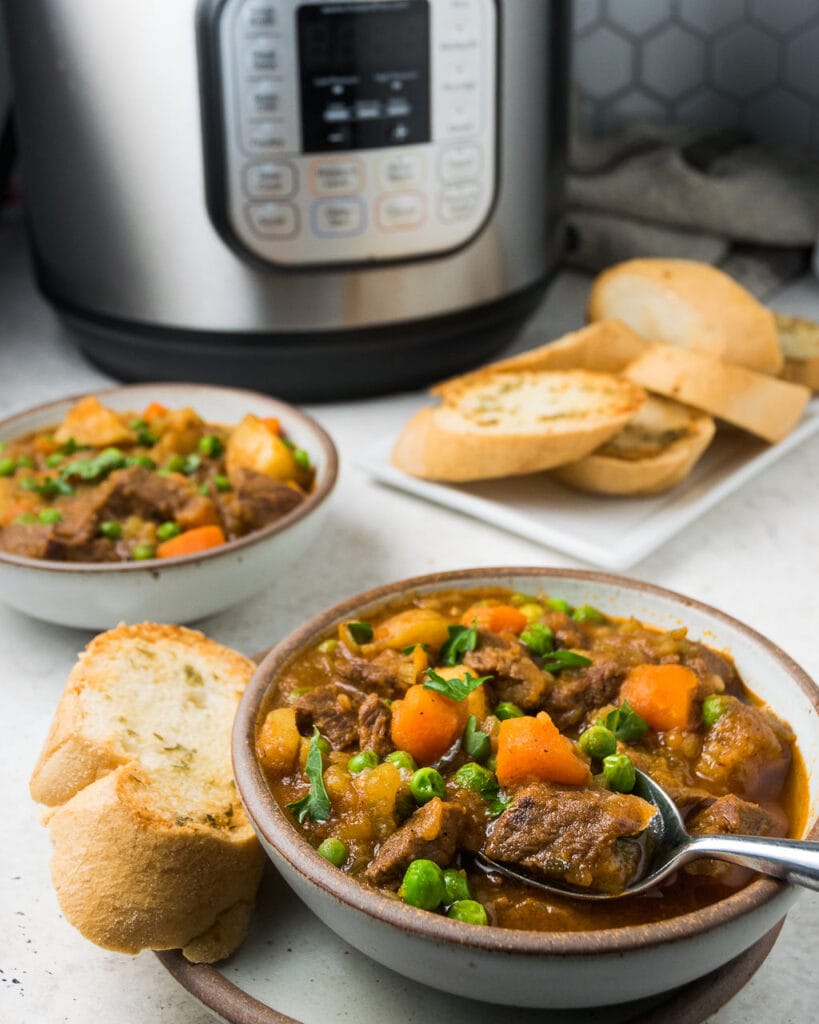 Two white bowls with beef stew with bread on the side on a white background with an instant pot in the back