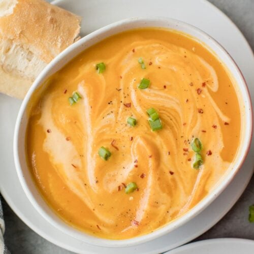 A bowl of Sweet Potato Soup on a white plate with bread off to the side
