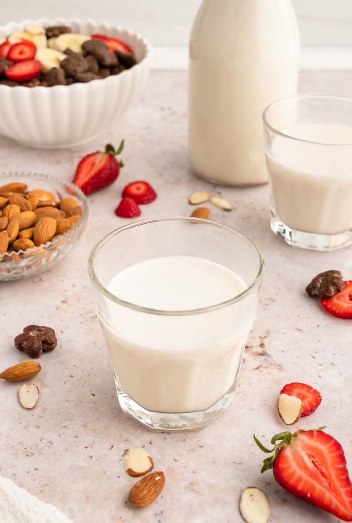 A clear glass of almond milk with a jar, glass, and strawberries in the background.