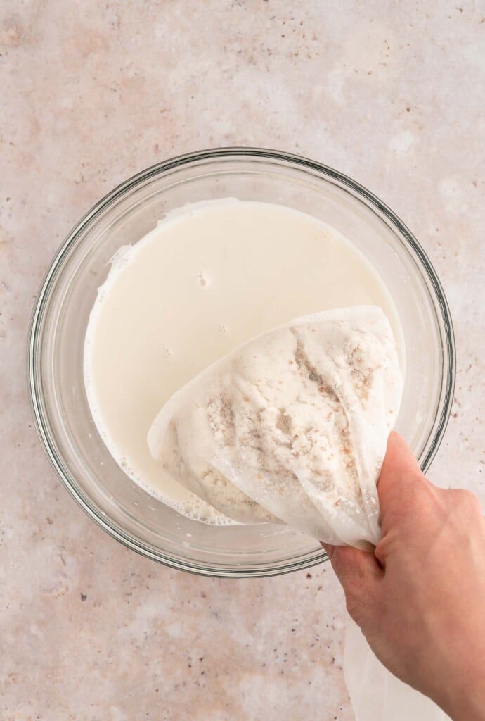 A hand holding a nut milk bag over a bowl of freshly squeezed almond milk.