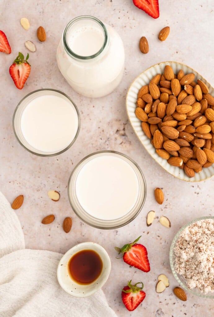 An overhead shot of two glasses of almond milk, a milk jar of almond milk and a plate full of almonds with strawberries on the side.