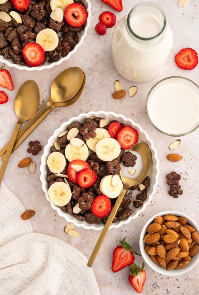 An overhead shot of a bowl of chocolate cereal, sliced strawberries and bananas with almond milk and almonds on the side.