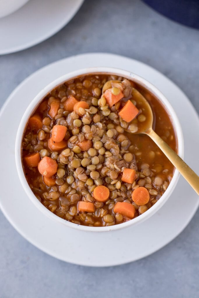 A bowl of easy lentil soup on a white plate with bread off to the side and a bite being taken out of it with a gold spoon
